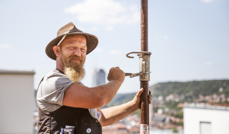 bärtiger Mitarbeiter auf Dach mit Zimmermannsstift hinter dem Ohr schraubt an einer Stützstange